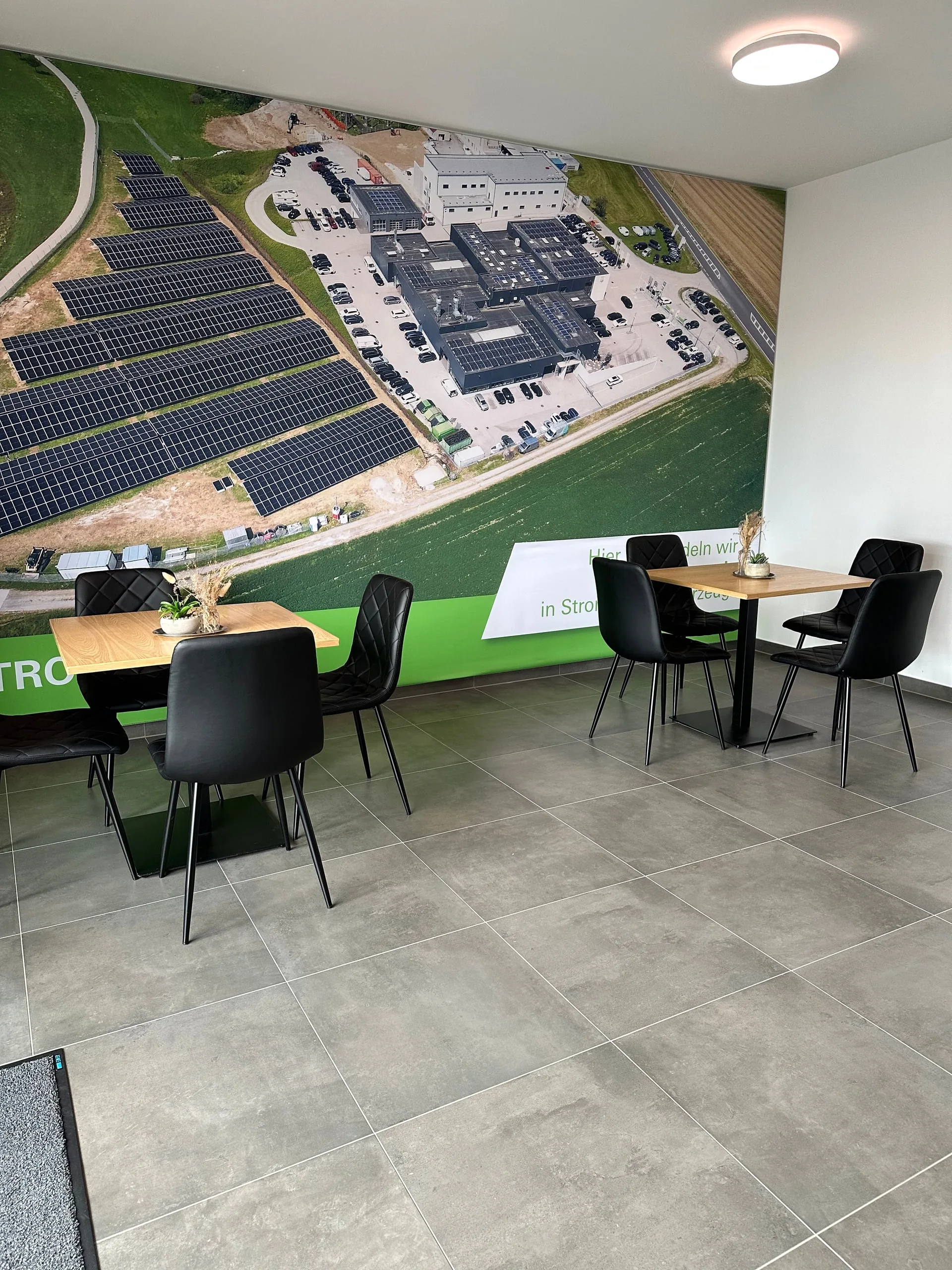 Lounge mit Tischen und großem Wandbild, das den Ladepark und das Photovoltaikfeld von oben zeigt
